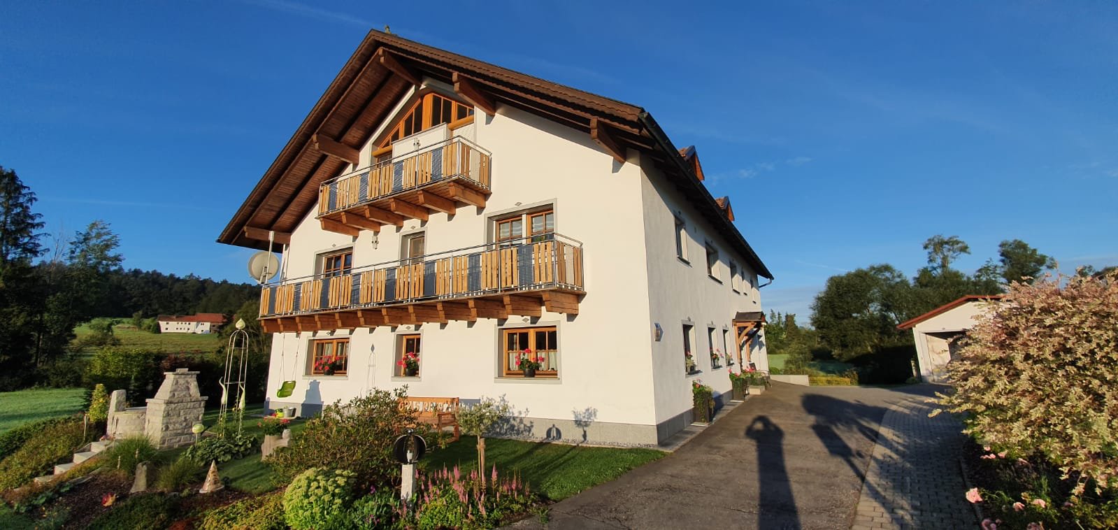 Das Bild zeigt ein Wohnhaus und davor einen Garten. Dahinter sind weitere Gebäude zusehen. Im Hintergrund ist ein blauer Himmel zu sehen